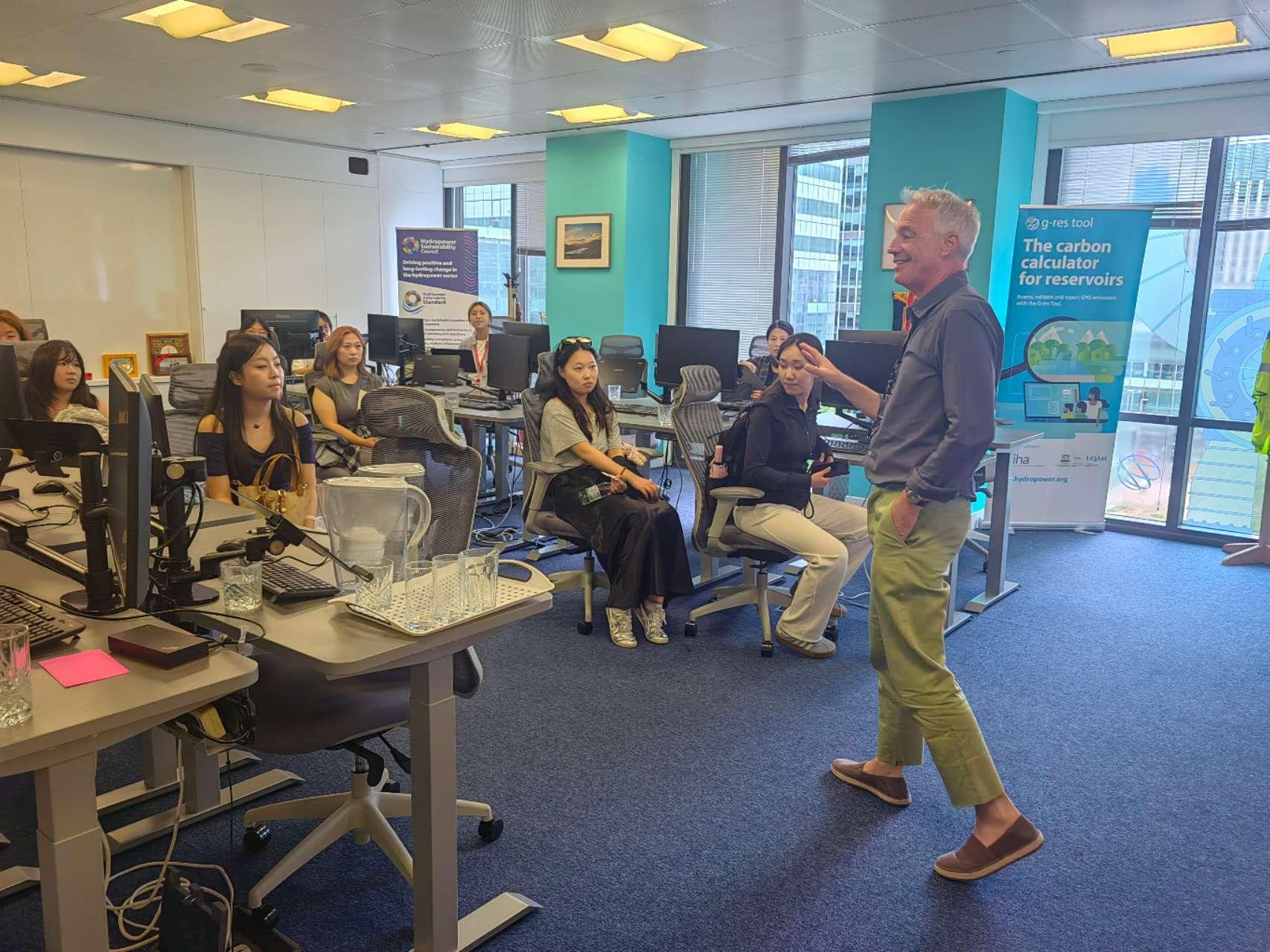 IHA CEO Eddie Rich speaks to seated youth delegates in an office beside sustainable hydropower displays.